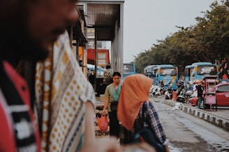 A busy street scene with people walking near a row of parked buses. A man is visible in the foreground with blurred focus, and a woman in a hijab is walking nearby. Motorcycles and additional figures are in the background, along with a street vendor cart.