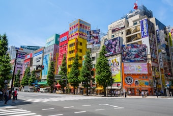 A vibrant urban street scene with tall buildings adorned with colorful advertisements and billboards, featuring anime characters and brand logos. Trees line the sidewalk, and pedestrians wait to cross the busy intersection. Vehicles are seen on the street, and the sky is clear and blue.