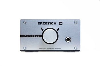 A silver headphone amplifier with a large central knob and various markings, including the brand name 'ERZETICH' and model name 'Perfidus'. There are four screws at each corner and an audio jack on the right side.