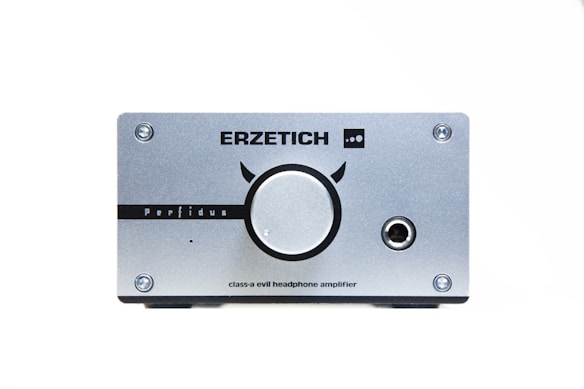 A silver headphone amplifier with a large central knob and various markings, including the brand name 'ERZETICH' and model name 'Perfidus'. There are four screws at each corner and an audio jack on the right side.