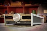 A sleek, high-end audio amplifier with a stylish wooden front panel and a large metallic control knob. The brand name 'Erzetich' and model 'Deimos' are visible. The background suggests a workshop or studio with shelves and various blurred objects.