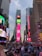 A bustling urban scene with a large crowd of people, tall buildings adorned with bright digital advertisements and billboards, and a clear, slightly cloudy sky in the background. The vibrant displays feature various brands and messages, including encouraging self-expression.