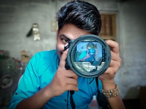 A person with dark hair holds a large camera. The lens captures their reflection, creating an interesting mirror image effect. They are wearing a bright blue shirt and a bracelet. The background is blurred, but appears to be an indoor setting.