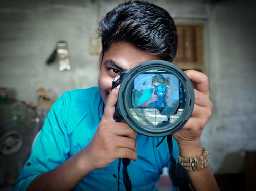 A person with dark hair holds a large camera. The lens captures their reflection, creating an interesting mirror image effect. They are wearing a bright blue shirt and a bracelet. The background is blurred, but appears to be an indoor setting.
