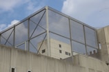 A modern building with a geometric design features a large metal mesh structure on the rooftop. The structure has a grid-like pattern, with rectangular glass panels reflecting the sky. The building facade shows a combination of concrete and glass elements.