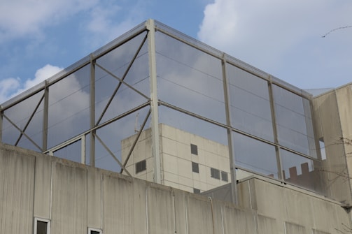 A modern building with a geometric design features a large metal mesh structure on the rooftop. The structure has a grid-like pattern, with rectangular glass panels reflecting the sky. The building facade shows a combination of concrete and glass elements.