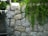 A natural stone wall constructed from large, unevenly shaped grey rocks. Lush green foliage cascades down the wall, contrasted by nearby green plants and trees.