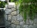A natural stone wall constructed from large, unevenly shaped grey rocks. Lush green foliage cascades down the wall, contrasted by nearby green plants and trees.