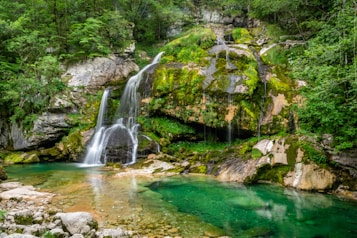 A serene waterfall cascades over moss-covered rocks into a clear turquoise pool, surrounded by lush green foliage and forested cliffs. The tranquil scene is framed by rugged terrain with a mix of rock formations and vibrant plant life, offering a picturesque representation of untouched natural beauty.