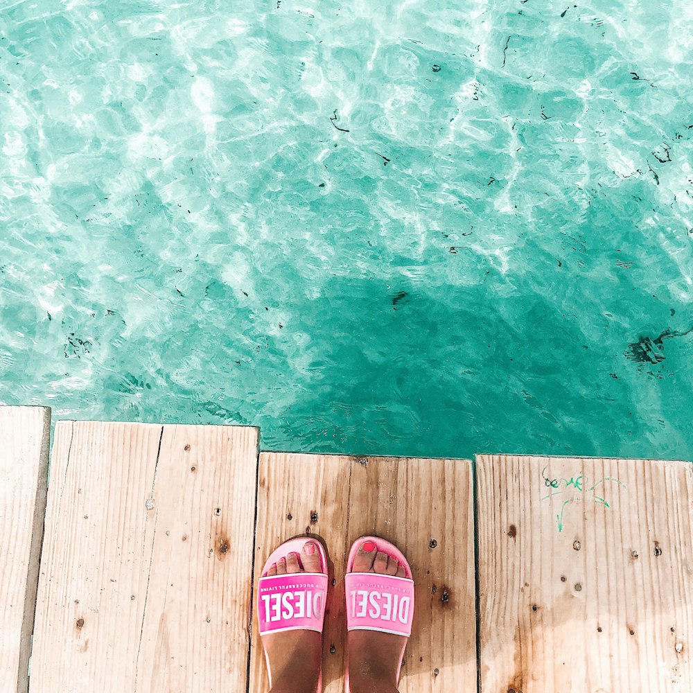 Frau trägt rosa Pantoffeln, die auf dem Dock vor klarem Wasser stehen