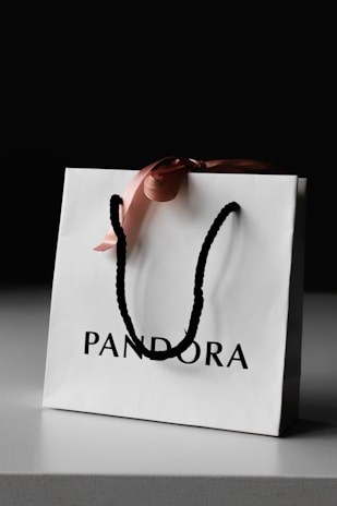 A white paper shopping bag with black handles sits on a gray surface. The bag is adorned with a pink ribbon tied in a bow and has the word 'PANDORA' printed on it in bold black letters.