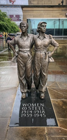 A bronze statue of two women in work attire, standing side by side with arms around each other, on a rainy pavement. The base reads 'Women of Steel 1914-1918 1939-1945'. The background shows a museum building with glass windows and some people walking in the rain.