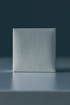 A minimalist photo featuring a square box with a soft texture and the word 'PANDORA' embossed on the front. The box is centered against a muted dark background and rests on a similarly textured surface. The overall aesthetic is clean and elegant.