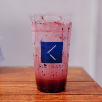 A plastic cup filled with a pink drink with visible black specks and a dark section at the bottom; the cup features a blue Kemenady logo with a white letter K. It is placed on a wooden surface with a light, blurred background.