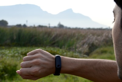A person checks a smart device on their wrist while standing in a natural setting. The background includes grassy fields and distant hills under a clear sky.