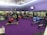 A gym interior with several people engaging in various physical exercises. The room has purple flooring and walls, and is equipped with gym equipment like rowing machines, plyo soft boxes, weights, and stability balls. There are mirrors on the walls and a TV screen displaying a video. Some individuals are performing bodyweight exercises, while others are using equipment. The lighting is bright, and the atmosphere appears active and focused.