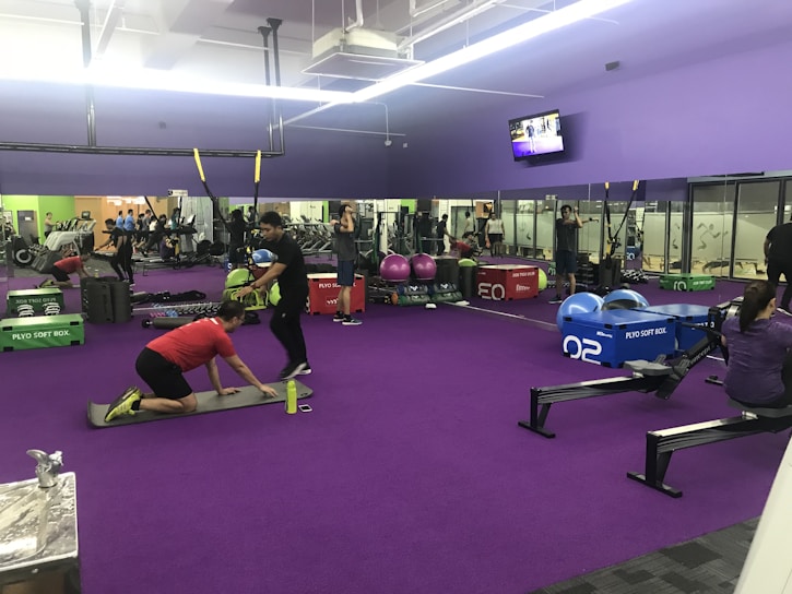 A gym interior with several people engaging in various physical exercises. The room has purple flooring and walls, and is equipped with gym equipment like rowing machines, plyo soft boxes, weights, and stability balls. There are mirrors on the walls and a TV screen displaying a video. Some individuals are performing bodyweight exercises, while others are using equipment. The lighting is bright, and the atmosphere appears active and focused.
