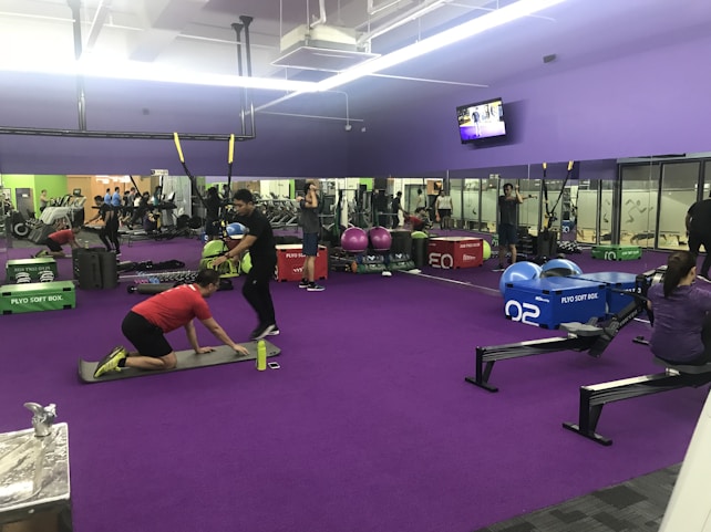 A gym interior with several people engaging in various physical exercises. The room has purple flooring and walls, and is equipped with gym equipment like rowing machines, plyo soft boxes, weights, and stability balls. There are mirrors on the walls and a TV screen displaying a video. Some individuals are performing bodyweight exercises, while others are using equipment. The lighting is bright, and the atmosphere appears active and focused.