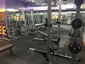 A well-equipped gym with various weightlifting equipment including benches, barbells, dumbbells, and weight racks. There are mirrors on the walls and rubber flooring. The lighting is bright yet soft, and the gym appears clean and organized.