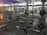 A well-equipped gym with various weightlifting equipment including benches, barbells, dumbbells, and weight racks. There are mirrors on the walls and rubber flooring. The lighting is bright yet soft, and the gym appears clean and organized.