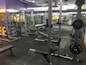 A well-equipped gym with various weightlifting equipment including benches, barbells, dumbbells, and weight racks. There are mirrors on the walls and rubber flooring. The lighting is bright yet soft, and the gym appears clean and organized.