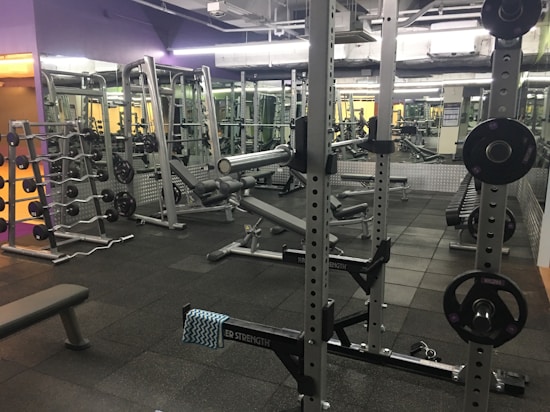A well-equipped gym with various weightlifting equipment including benches, barbells, dumbbells, and weight racks. There are mirrors on the walls and rubber flooring. The lighting is bright yet soft, and the gym appears clean and organized.