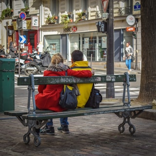 A couple is sitting on a park bench, with arms around each other's shoulders. They are facing away from the camera, with a street view in the background featuring parked scooters and a visible HSBC bank branch. The couple is dressed in bright clothing, with the woman wearing a red coat and the man in a yellow one. Graffiti is visible on the bench, and a few pedestrians can be seen in the background.
