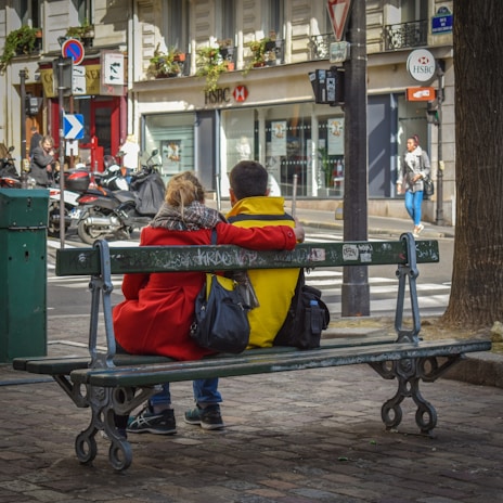 A couple is sitting on a park bench, with arms around each other's shoulders. They are facing away from the camera, with a street view in the background featuring parked scooters and a visible HSBC bank branch. The couple is dressed in bright clothing, with the woman wearing a red coat and the man in a yellow one. Graffiti is visible on the bench, and a few pedestrians can be seen in the background.