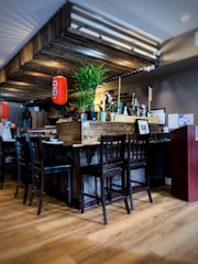 A cozy interior with wooden furniture and flooring, featuring a central table surrounded by chairs. A vibrant red lantern hangs above, contrasting with green potted plants. Various bottles and decorative items are arranged on the table, adding to a homely atmosphere.