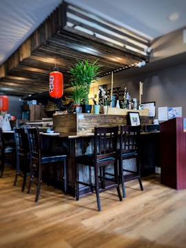 A cozy interior with wooden furniture and flooring, featuring a central table surrounded by chairs. A vibrant red lantern hangs above, contrasting with green potted plants. Various bottles and decorative items are arranged on the table, adding to a homely atmosphere.