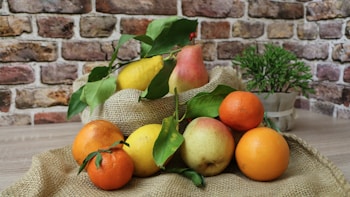 A rustic scene featuring a variety of fresh fruits, including pears, lemons, oranges, and mandarins, arranged on a piece of burlap fabric. The fruits have vibrant colors with green leaves attached. In the background, there is a textured brick wall and a small potted plant, adding to the natural and earthy setting.