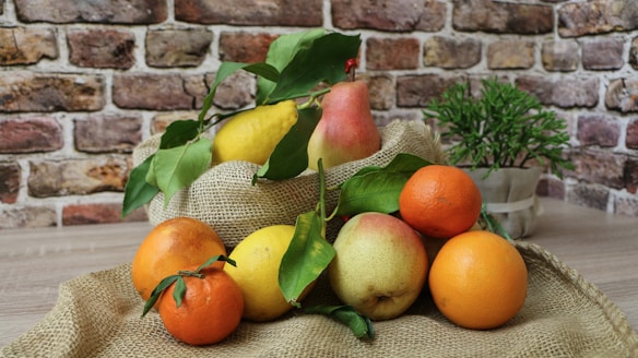 A rustic scene featuring a variety of fresh fruits, including pears, lemons, oranges, and mandarins, arranged on a piece of burlap fabric. The fruits have vibrant colors with green leaves attached. In the background, there is a textured brick wall and a small potted plant, adding to the natural and earthy setting.