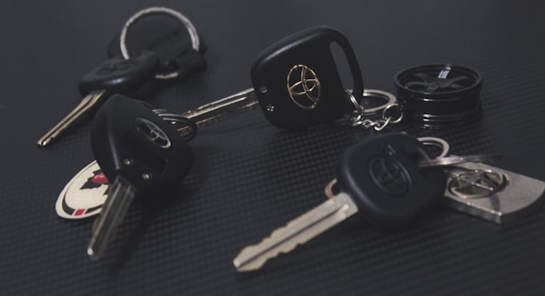 A collection of car keys with black key fobs displaying a logo, laid out on a dark textured surface. Some keys are attached to keychains, and one includes a round object resembling a miniature tire.