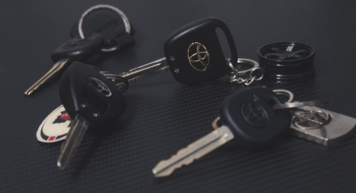 A collection of car keys with black key fobs displaying a logo, laid out on a dark textured surface. Some keys are attached to keychains, and one includes a round object resembling a miniature tire.