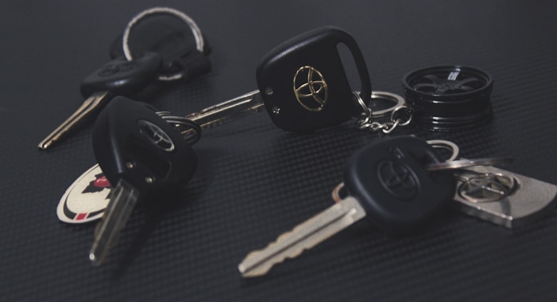 A collection of car keys with black key fobs displaying a logo, laid out on a dark textured surface. Some keys are attached to keychains, and one includes a round object resembling a miniature tire.