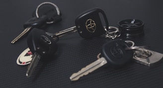 A collection of car keys with black key fobs displaying a logo, laid out on a dark textured surface. Some keys are attached to keychains, and one includes a round object resembling a miniature tire.
