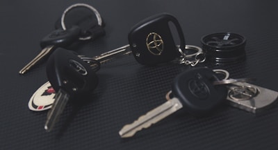 A collection of car keys with black key fobs displaying a logo, laid out on a dark textured surface. Some keys are attached to keychains, and one includes a round object resembling a miniature tire.