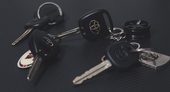 A collection of car keys with black key fobs displaying a logo, laid out on a dark textured surface. Some keys are attached to keychains, and one includes a round object resembling a miniature tire.