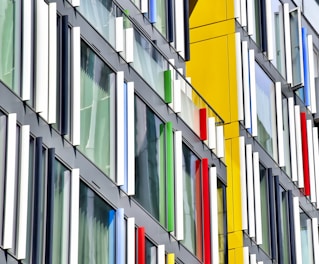 A modern building facade features large glass windows with colorful vertical panels in blue, red, white, and green. The windows are set in a sleek, dark frame with sections of bright yellow wall. The design creates a dynamic and playful pattern.