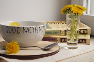 A white bowl with the text 'Good Morning' is placed on a wooden surface. Beside it, there is a small vase containing a few bright yellow dandelions. Some books with Cyrillic titles are stacked next to the vase. A spoon rests on the surface near the bowl.