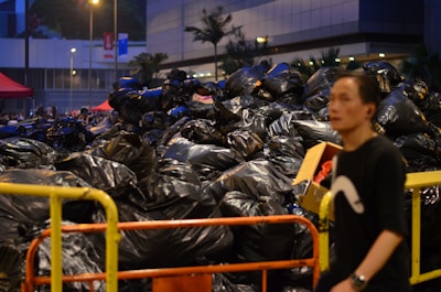 A large pile of black garbage bags fills the majority of the scene, likely containing various waste items. To the right, there is a person in a black shirt with a white design, walking past the pile. There are some boxes among the garbage bags, and portable barriers are visible, possibly for containing the trash. The background includes a building, street lights, and a tent, indicating an urban setting.