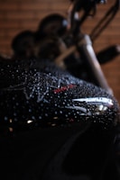The image captures a close-up view of a motorcycle, specifically its black metallic surface covered in water droplets. The background is blurry, highlighting the shiny surface with red and white accents.