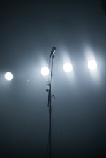 A solitary microphone stands on a dimly lit stage with bright spotlights shining in the background, creating a sense of anticipation.