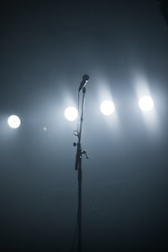 A solitary microphone stands on a dimly lit stage with bright spotlights shining in the background, creating a sense of anticipation.