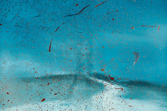 A close-up view of a surface covered with numerous tiny specks and splashes of brownish-red paint against a bright turquoise background. The texture appears irregular, with a blend of smooth and slightly gritty areas, creating an abstract pattern.