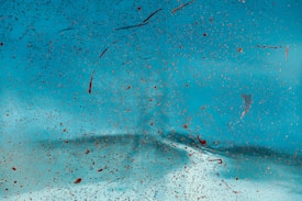 A close-up view of a surface covered with numerous tiny specks and splashes of brownish-red paint against a bright turquoise background. The texture appears irregular, with a blend of smooth and slightly gritty areas, creating an abstract pattern.