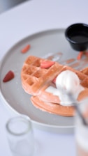 Close-up of a stack of golden waffles topped with fresh strawberries, whipped cream, and a drizzle of chocolate syrup.