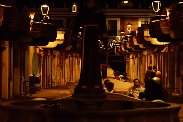 A dimly lit street with a series of hanging lanterns casting a warm glow on the buildings lining the path. People are gathered in small groups, engaging in conversation. The scene exudes a cozy, intimate atmosphere, with visible shadows enhancing the mood.