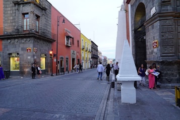 A cobblestone street lined with colorful historic buildings, featuring orange and yellow facades. People are walking and gathering in groups along the sidewalk. A corner of a grand stone building with ornate detailing is visible, and various pedestrians pass by in casual outfits. Traffic lights and street lamps are present, contributing to the urban atmosphere.