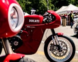 A striking red Ducati motorcycle is prominently displayed, showcasing its sleek design and engineering. The background features a blurred outdoor setting with tents and people, suggesting a bike show or event atmosphere.